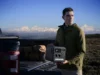 A man in an olive jacket holds a portable solar generator beside a pickup truck and camping gear, with snowy mountains beyond; rugged, contemplative mood.