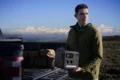 A man in an olive jacket holds a portable solar generator beside a pickup truck and camping gear, with snowy mountains beyond; rugged, contemplative mood.