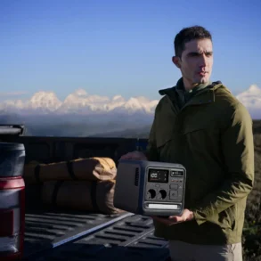 A man in an olive jacket holds a portable solar generator beside a pickup truck and camping gear, with snowy mountains beyond; rugged, contemplative mood.