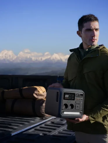A man in an olive jacket holds a portable solar generator beside a pickup truck and camping gear, with snowy mountains beyond; rugged, contemplative mood.