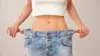 A person pulls oversized blue jeans away from their waist, showing weight loss; the loose fit highlights a slim midsection against a plain background.