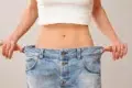 A person pulls oversized blue jeans away from their waist, showing weight loss; the loose fit highlights a slim midsection against a plain background.