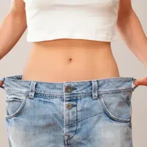 A person pulls oversized blue jeans away from their waist, showing weight loss; the loose fit highlights a slim midsection against a plain background.