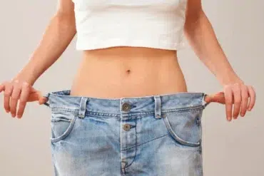 A person pulls oversized blue jeans away from their waist, showing weight loss; the loose fit highlights a slim midsection against a plain background.