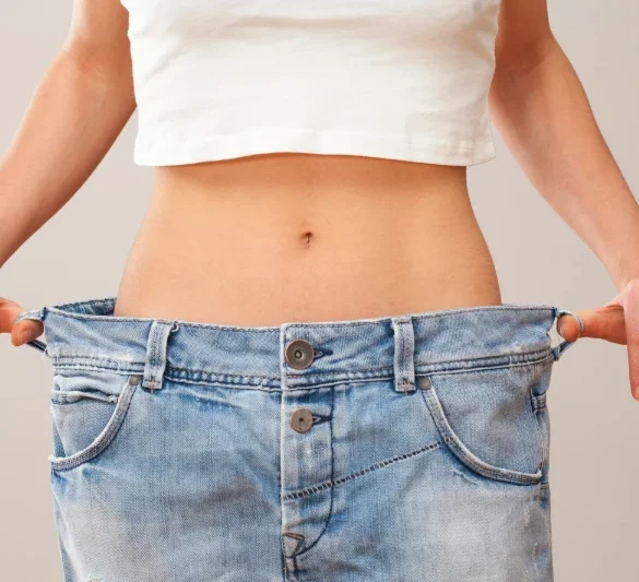 A person pulls oversized blue jeans away from their waist, showing weight loss; the loose fit highlights a slim midsection against a plain background.