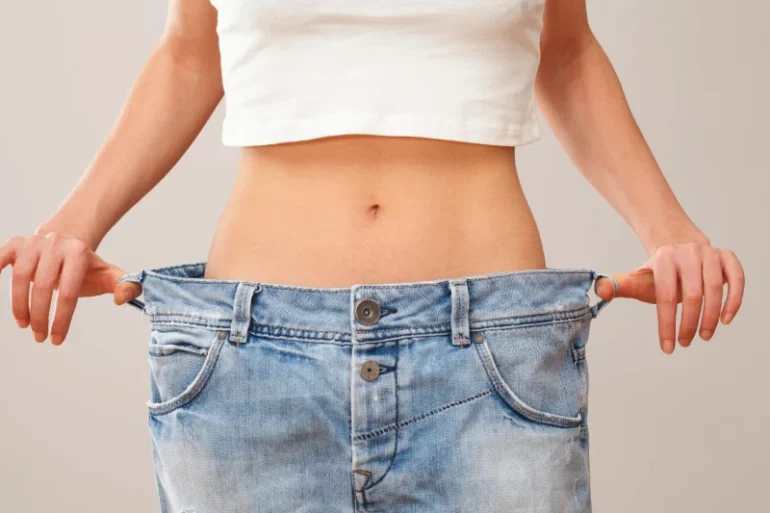 A person pulls oversized blue jeans away from their waist, showing weight loss; the loose fit highlights a slim midsection against a plain background.