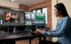 A focused video editor works at dual monitors, adjusting color and editing guitar performance footage in a bright studio with brick walls.