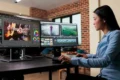 A focused video editor works at dual monitors, adjusting color and editing guitar performance footage in a bright studio with brick walls.